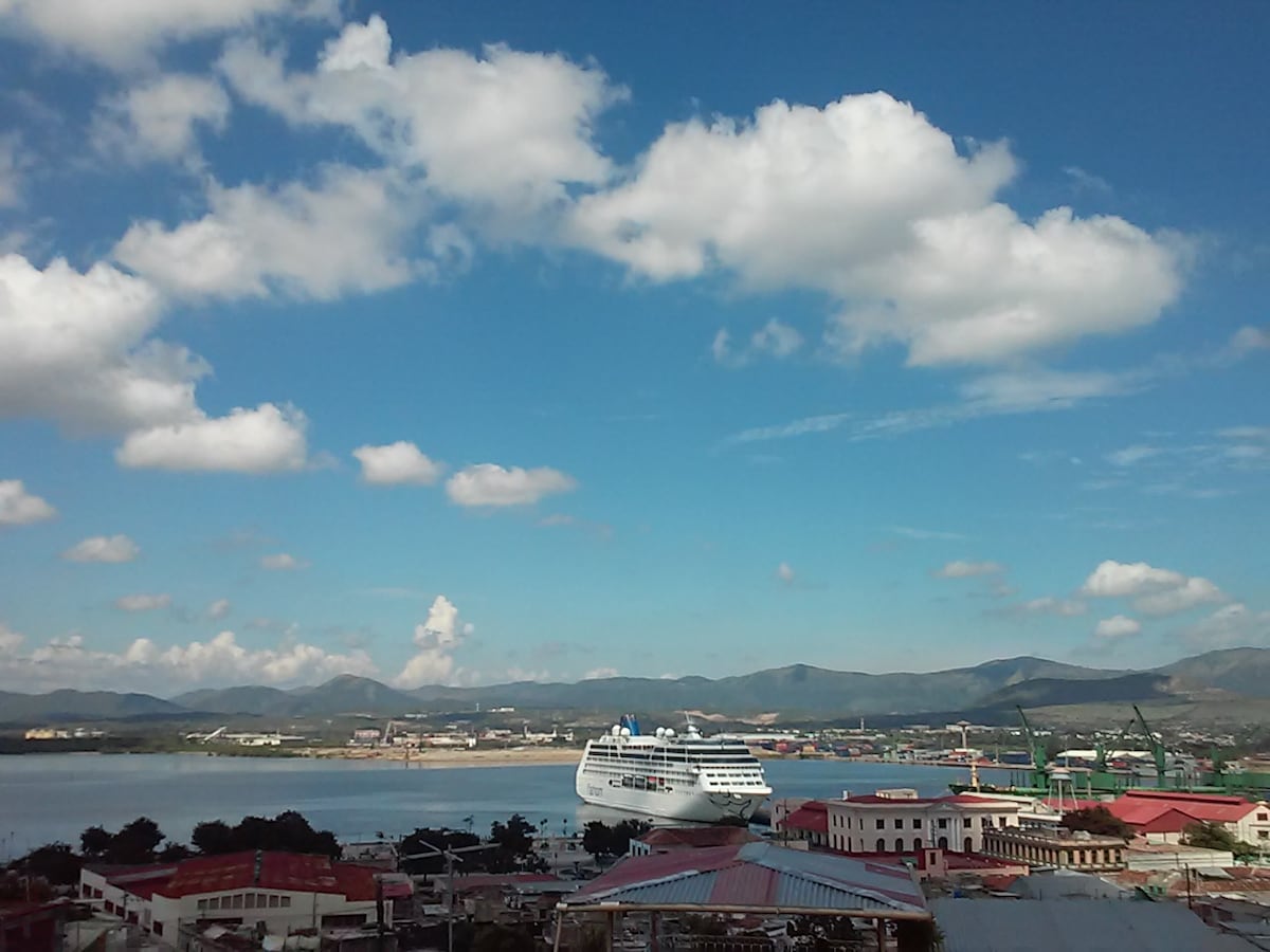 A panoramic view of a harbor showcases a large cruise ship moored in the water. The background is filled with distant mountains and industrial structures, under a bright blue sky dotted with white clouds.