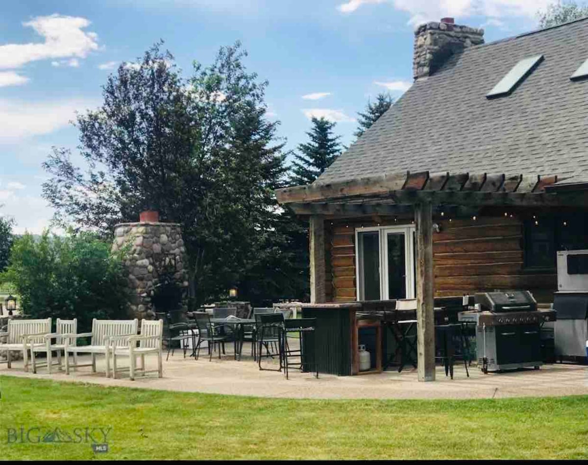 An outdoor patio area features a stone fireplace and two barbecue grills, surrounded by multiple seating options. The lodge's rustic exterior is complemented by lush green landscaping, creating an inviting space for gatherings and outdoor dining.