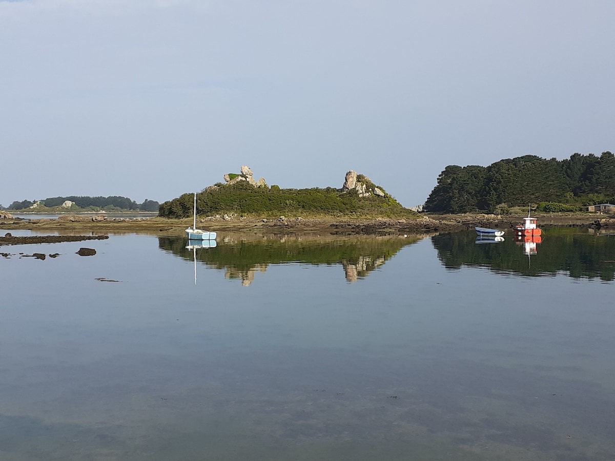A serene coastal scene is depicted, showcasing calm waters that reflect the surrounding greenery and rocky formations. Two small boats, one blue and one red, are anchored nearby, contributing to the tranquil atmosphere of the landscape.