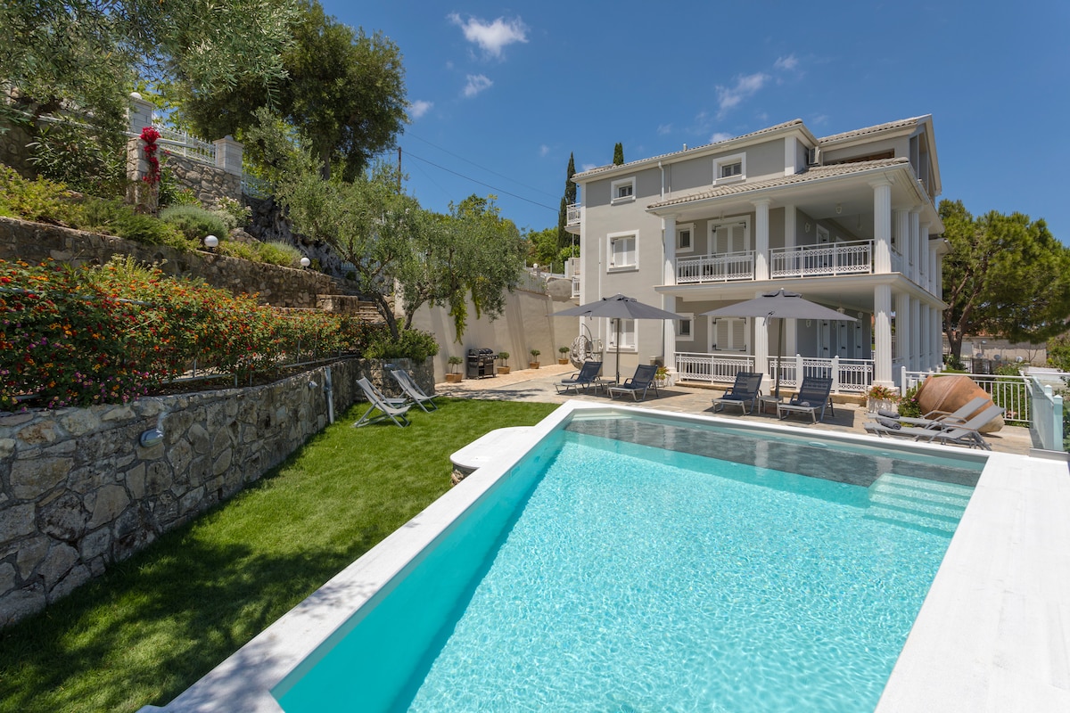 A well-maintained outdoor area features a private swimming pool surrounded by sun loungers under large umbrellas. The three-story villa displays a modern design with balconies on each level, and lush greenery adds to the serene atmosphere.