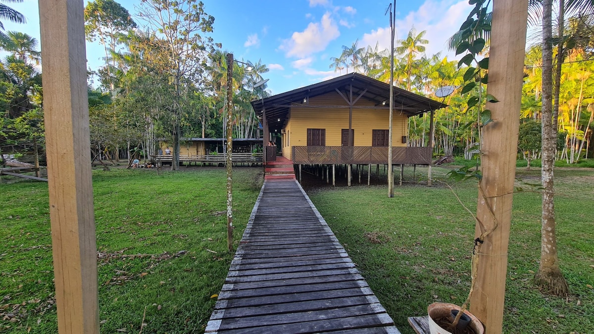 A wooden path leads towards a raised yellow structure surrounded by lush greenery. The building features a wide porch with evenly spaced windows, blending with the natural landscape. Trees and open spaces frame the scene, highlighting the tranquility of the environment.