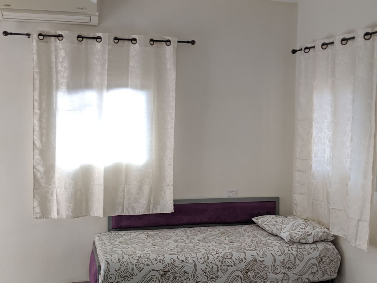 A minimalist sleeping area features a single bed with a patterned duvet and pillow. Natural light filters through sheer white curtains, illuminating the space. A neutral wall color enhances the bright atmosphere, and an air conditioning unit is mounted above the bed.