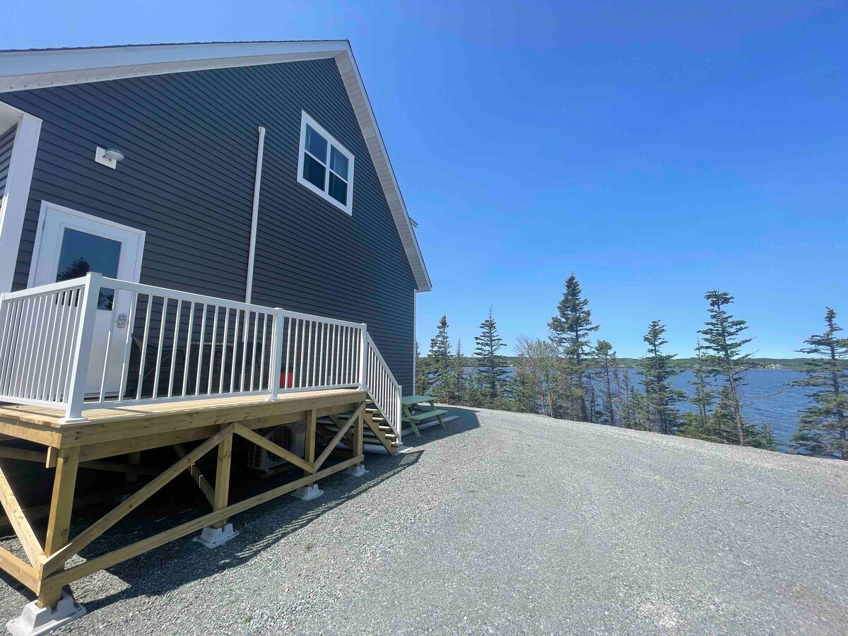 The exterior of the beach loft is showcased, featuring a grey siding with a large porch. A set of stairs leads to the entrance, framed by a gravel driveway. The view of the water and surrounding trees is visible in the background under a clear blue sky.