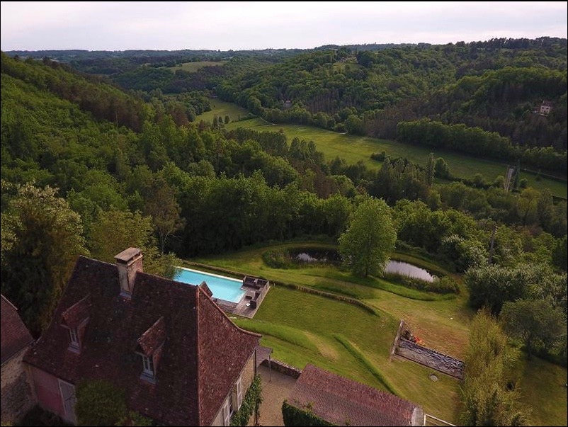 Aerial view of a spacious property with lush green surrounding hills. A sleek swimming pool is centrally located within manicured lawns, complemented by a tranquil pond. The authentic stone buildings and trees create a serene environment, illustrating the scenic countryside landscape.