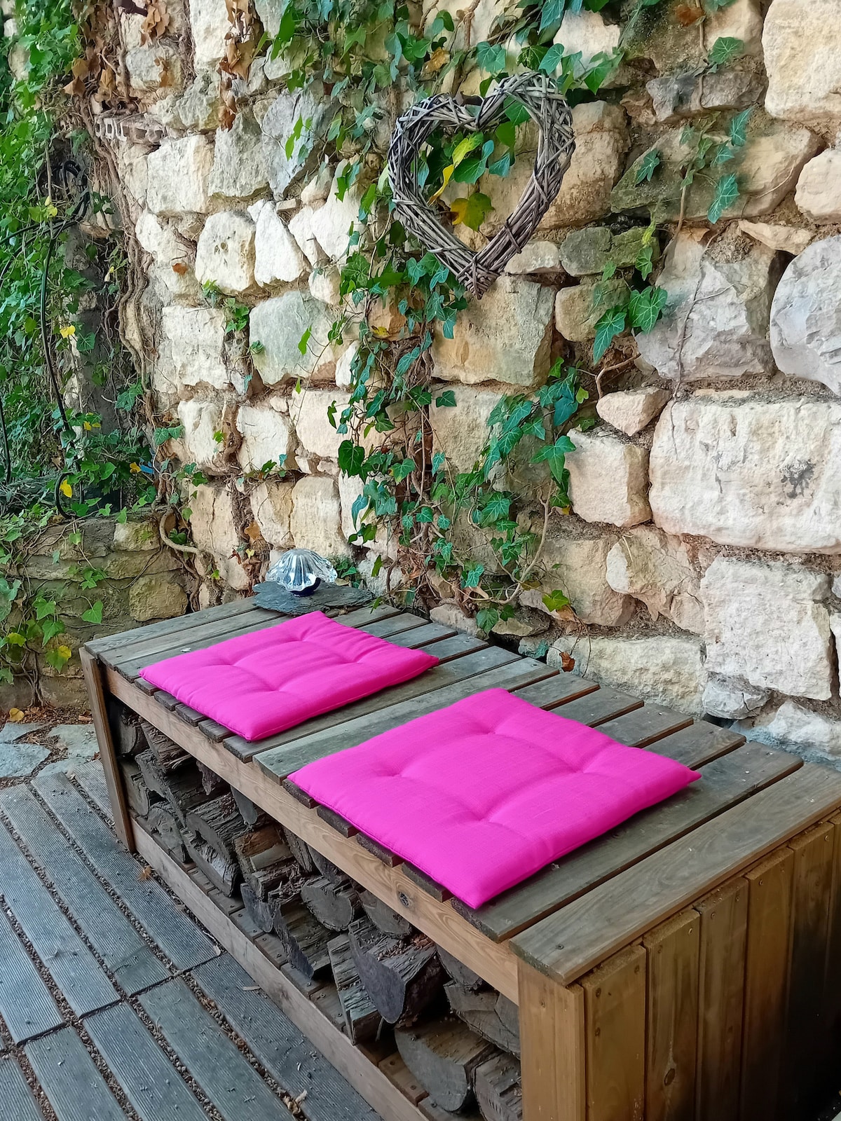 A wooden bench with bright pink cushions is placed against a weathered stone wall, surrounded by greenery. A heart-shaped decorative element hangs above, complementing the natural setting. The floor features wooden planks, contributing to a rustic atmosphere.