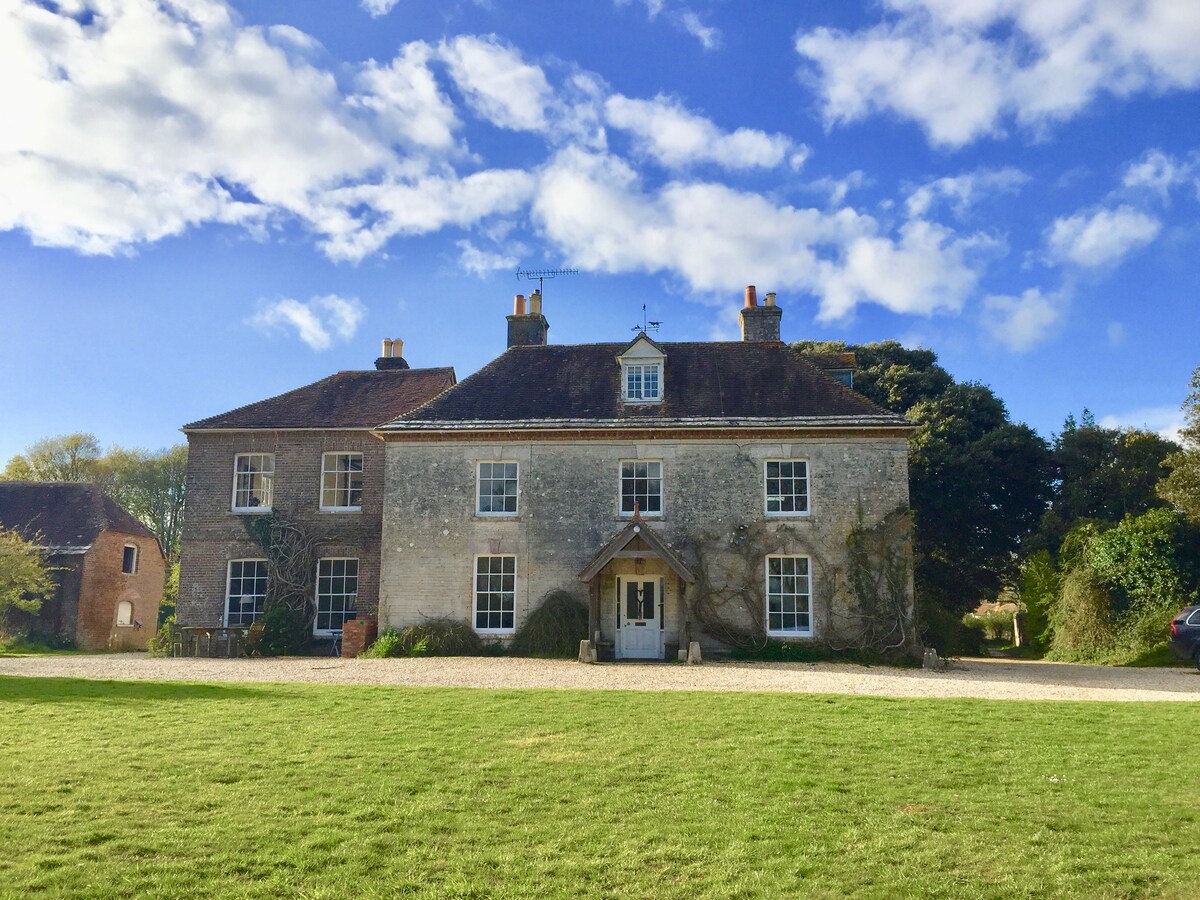 An 18th-century stone house is framed by a well-manicured lawn, with various trees surrounding the property. A front entrance, featuring a small porch, is flanked by multiple windows reflecting the sky. The bright blue sky is dotted with clouds, enhancing the serene setting.