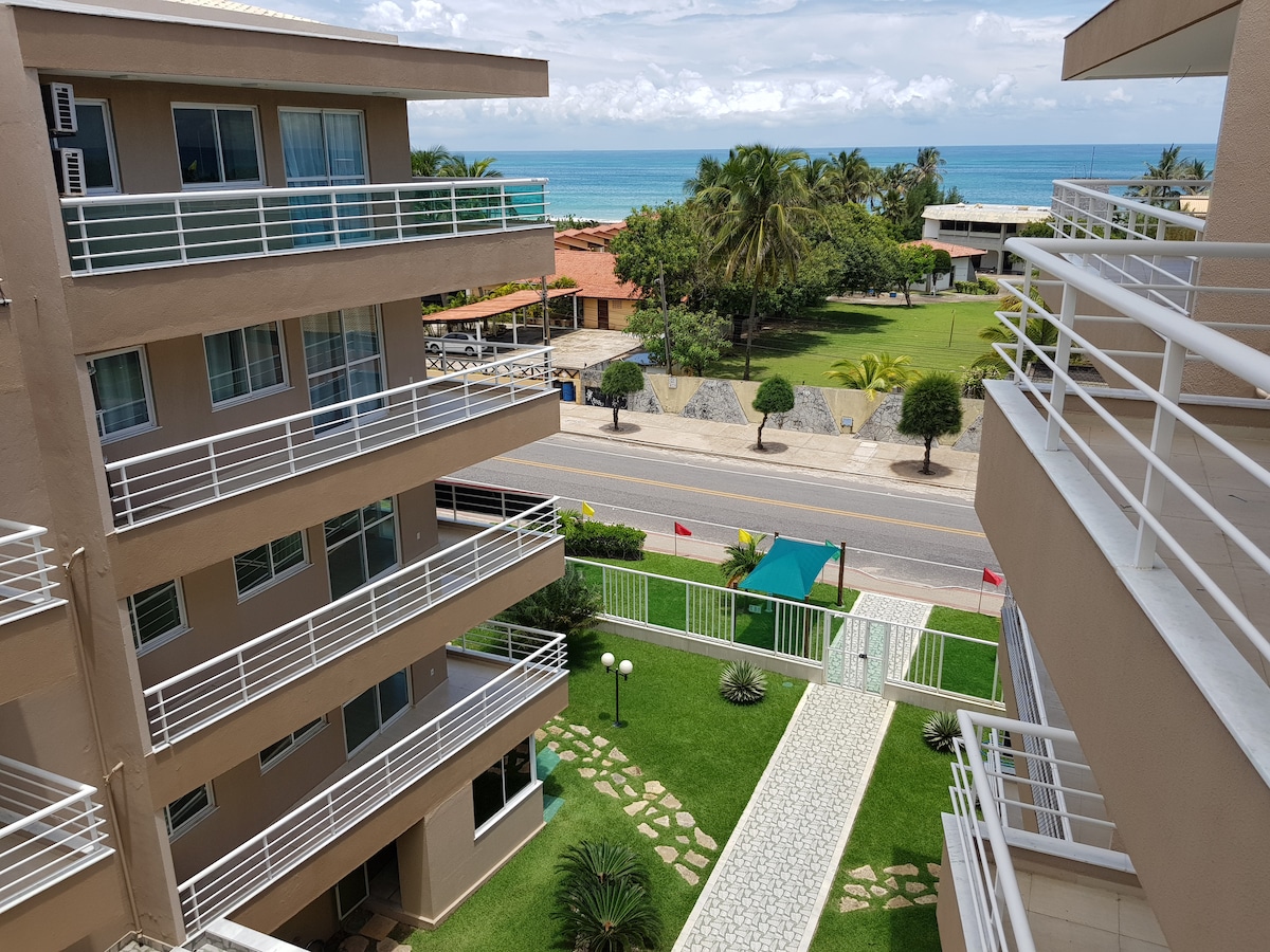 A view from a balcony showcases the landscaped grounds below, featuring a pathway leading to a green area. In the distance, the ocean glimmers under a partly cloudy sky, with palm trees swaying gently. Nearby structures are visible, contributing to a relaxed atmosphere.