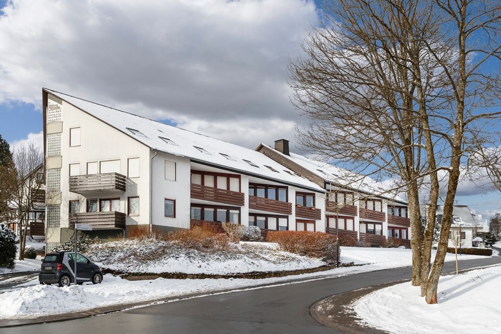 A modern apartment building is situated on a snowy landscape, featuring a sloping roof and multiple balconies. The exterior is predominantly white, complemented by wooden accents. A parked car can be seen near the entrance, with trees lining the quiet street.