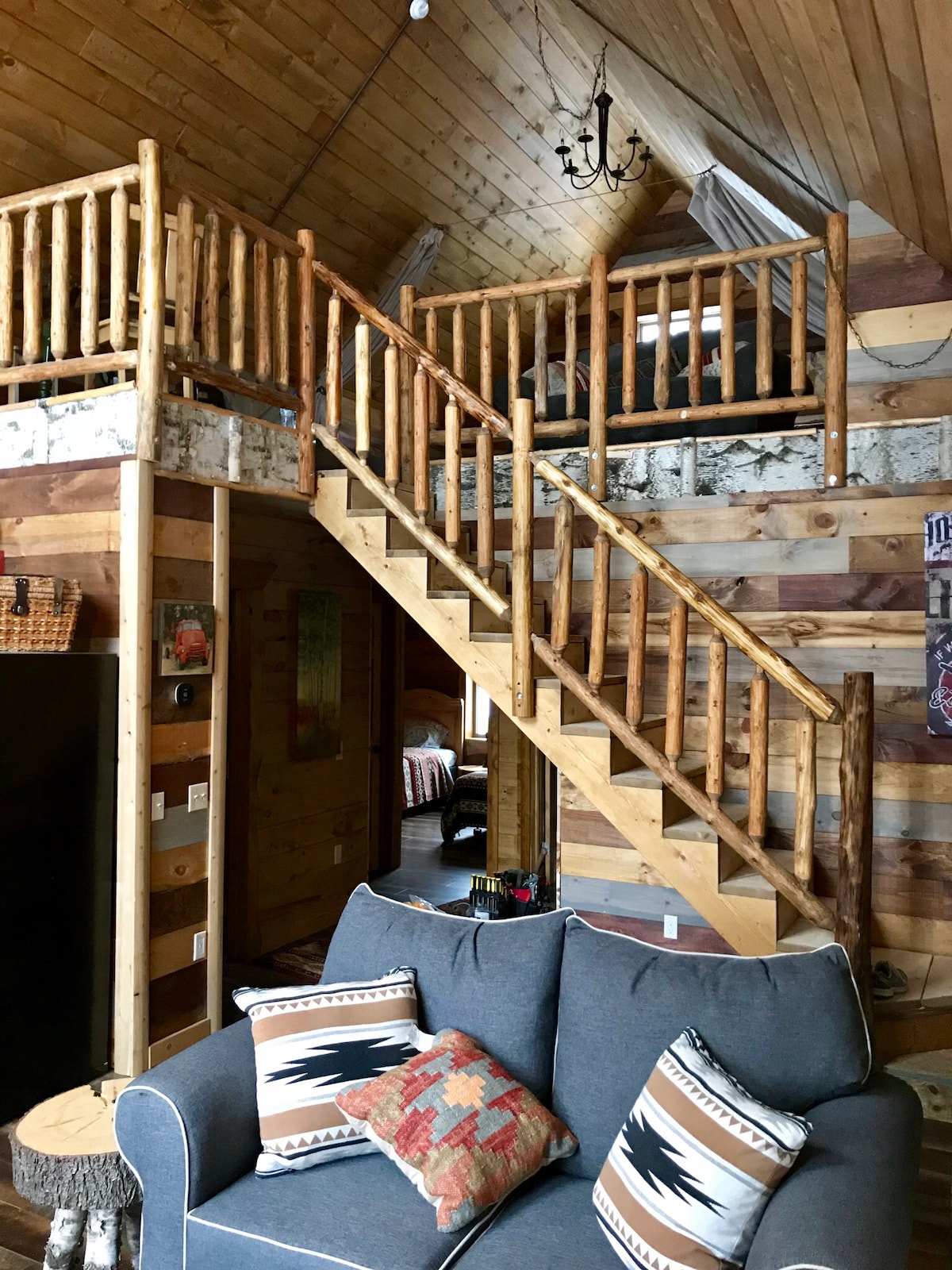 A wooden staircase with log railings leads to a loft area, showcasing a rustic design. The interior features natural wood paneling, adding warmth to the space. A comfortable gray sofa, adorned with decorative pillows, is situated in the foreground, inviting relaxation.