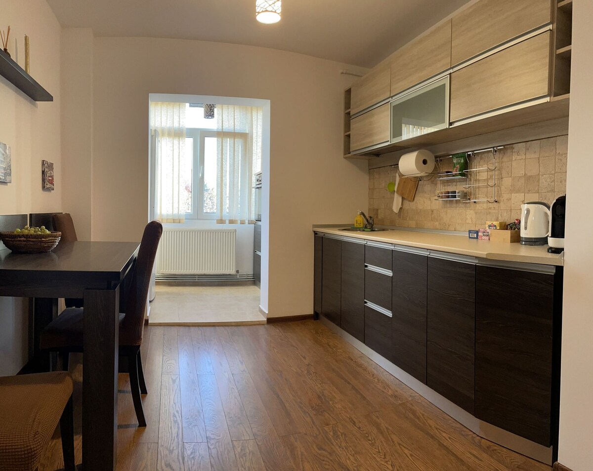 A spacious kitchen area is equipped with dark wood cabinetry and neutral-toned countertops. A wooden dining table is accompanied by four chairs, while a window allows natural light to fill the space. Decorative items and kitchen essentials are neatly arranged on the countertops.