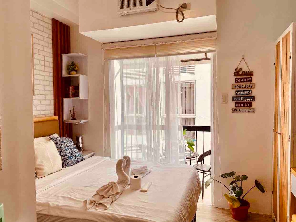A bright studio interior features a spacious bed adorned with decorative pillows. Natural light filters through sheer curtains, illuminating the room. A small balcony is visible, accompanied by a potted plant. Shelving units offer practical storage space, enhancing the overall functionality.