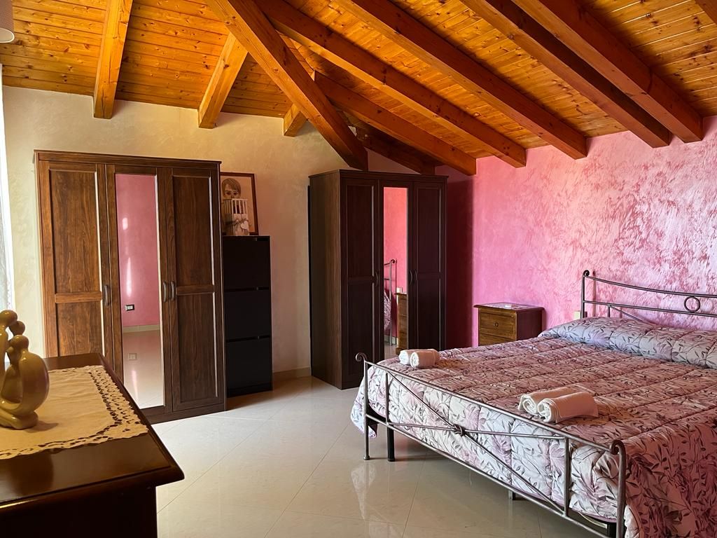 A spacious bedroom is presented with a wooden beamed ceiling and soft pink walls. A large bed with floral bedding is featured prominently. Storage is provided by a wardrobe and dresser, while natural light filters through the room, creating a serene environment.