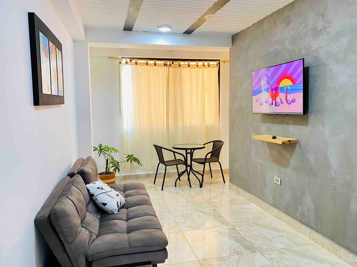 A modern living area is displayed with a gray sectional sofa and a round table with two chairs. Natural light filters through the curtains, creating a bright space. A wall-mounted television features a colorful image, while a small plant adds a touch of greenery.
