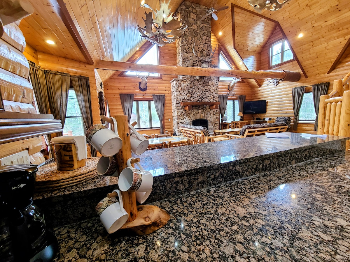 A spacious kitchen area features granite countertops and a coffee maker. A wooden mug holder displays several mugs. The background includes a living space with a tall stone fireplace and large windows that reveal views of the surrounding greenery. Log-style furniture adds to the rustic charm.