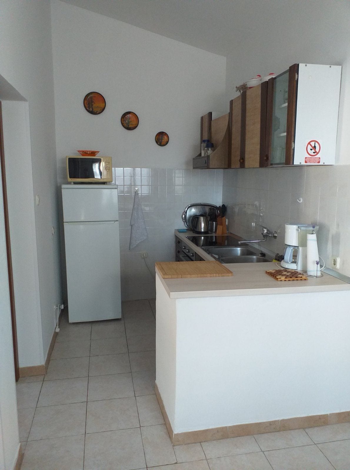 The kitchen area features modern appliances, including a refrigerator and a microwave. Wooden cabinets above the sink provide storage, while a countertop offers space for meal preparation. The tiled floor and light-colored walls contribute to a bright, functional cooking environment.