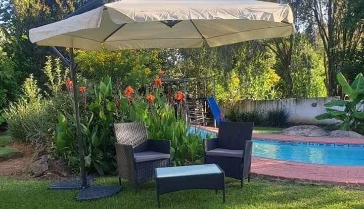 An outdoor seating area features two dark wicker chairs and a glass-topped table beneath a large umbrella. Lush greenery surrounds the space, and a swimming pool is visible in the background, complemented by vibrant flowers and a play structure.