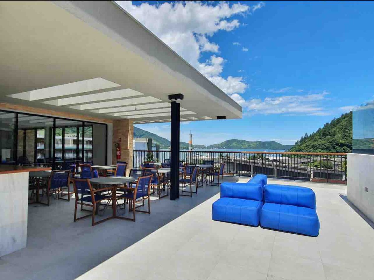 A spacious rooftop area is equipped with a seating arrangement of blue lounge furniture and tables surrounded by wooden chairs. Panoramic views of the mountains and surrounding landscape are visible in the background, under a clear blue sky with scattered clouds.