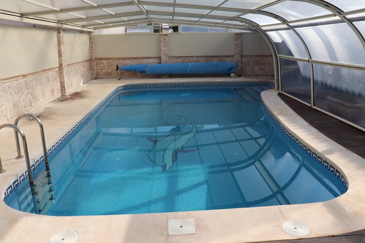 An indoor swimming pool is enclosed within a glass structure. The water is clear, revealing a mosaic design on the bottom. A ladder is positioned at one end for easy access, and a blue cover is seen rolled at the far side.