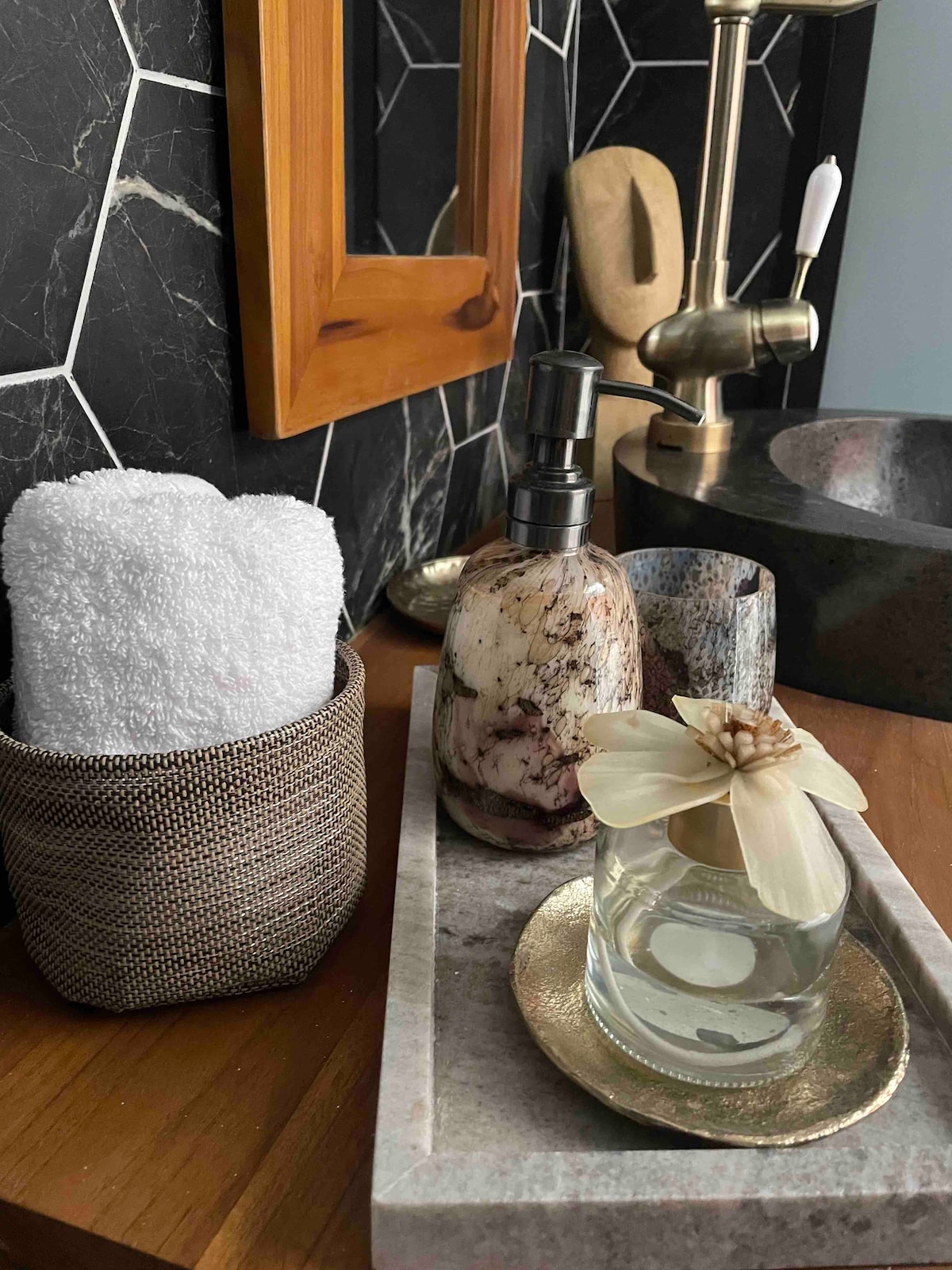 A neatly arranged vanity displays a woven basket containing rolled white towels. Various toiletries are showcased, including a decorative soap dispenser and a glass container holding a flower. A small tray with a metallic accent complements the natural textures present in the space.