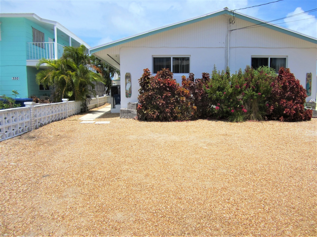 A spacious outdoor area is shown, featuring a gravel surface with clean landscaping. A lush garden with colorful foliage and shrubs borders the property, while clear blue skies and a glimpse of neighboring buildings can be seen in the background.