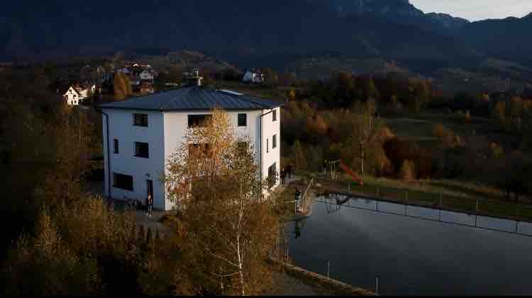 The villa features a modern hexagonal architectural design surrounded by autumn foliage. A serene pond reflects the building, offering a natural backdrop. The mountains are visible in the distance, completing the peaceful outdoor setting.