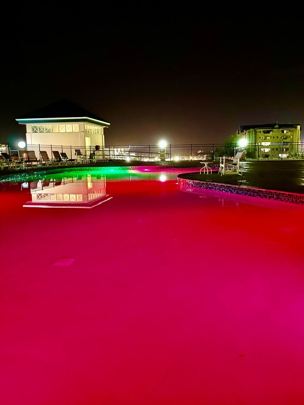 The pool area is shown at night, featuring vibrant colored lighting reflecting off the water's surface. Surrounding lounge chairs are illuminated by soft light, while a nearby building adds to the tranquil ambiance.