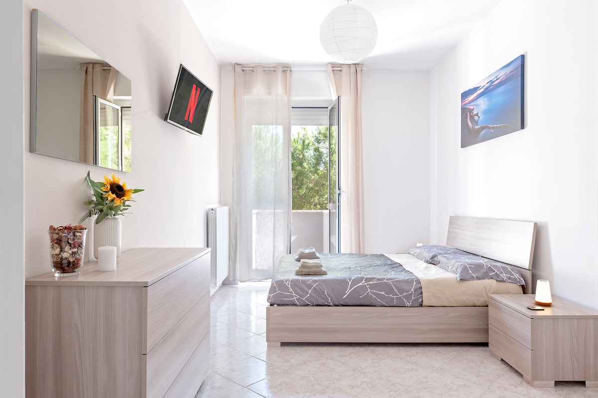 A spacious bedroom is filled with natural light, featuring a queen-size bed with neutral bedding. A large window opens to a balcony, while a dresser and bedside table provide functional storage. A decorated wall and a mounted television enhance the room's modern decor.