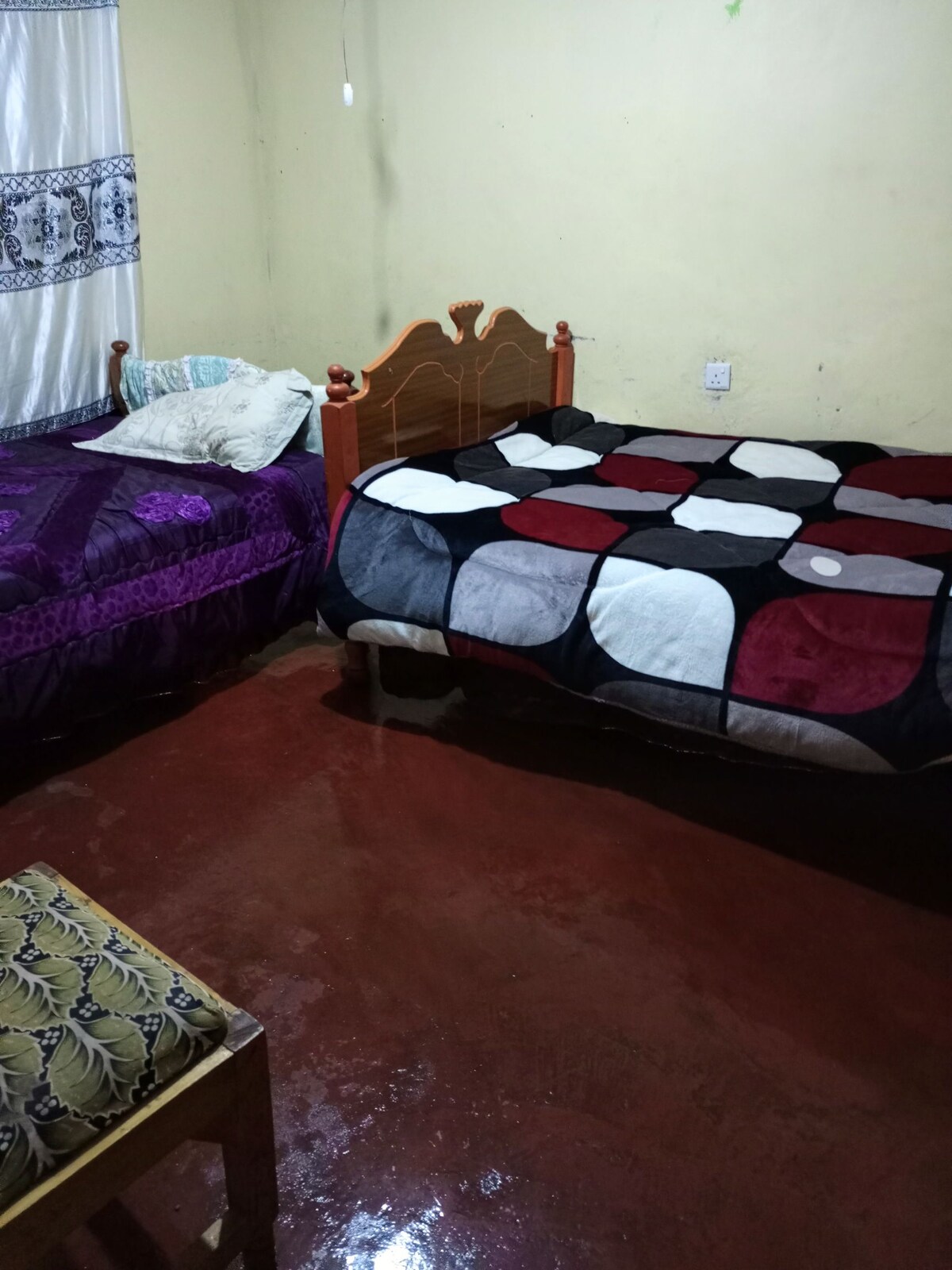 A simple bedroom is showcased with two beds, one with a purple coverlet and the other featuring a patterned blanket in red, black, and white. A small chair is positioned nearby. Natural light filters through a window draped with a white and blue curtain.