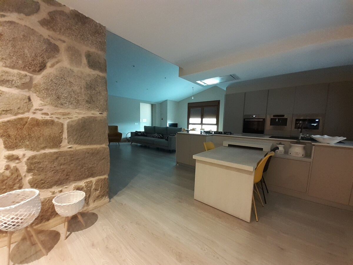 An open-concept kitchen and living area features a stone wall, enhancing the modern design. A light-colored countertop contrasts with sleek cabinetry. A seating area is visible in the background, complemented by soft natural light from large windows.