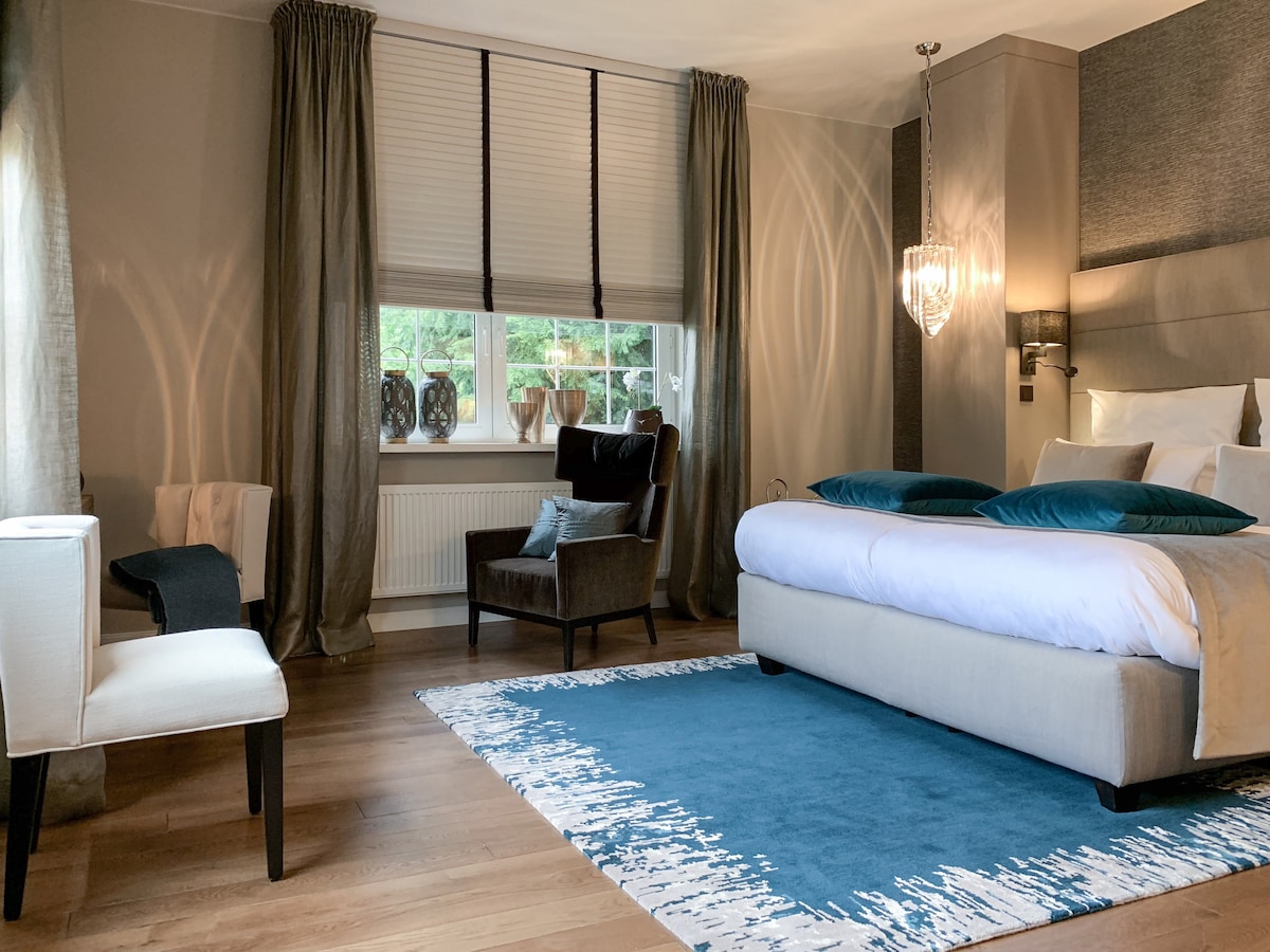 A spacious bedroom features a plush bed with decorative pillows and a soft area rug in shades of blue. Natural light filters through layered window treatments, which frame a view of outdoor greenery. A stylish armchair and a small accent chair complete the inviting space.