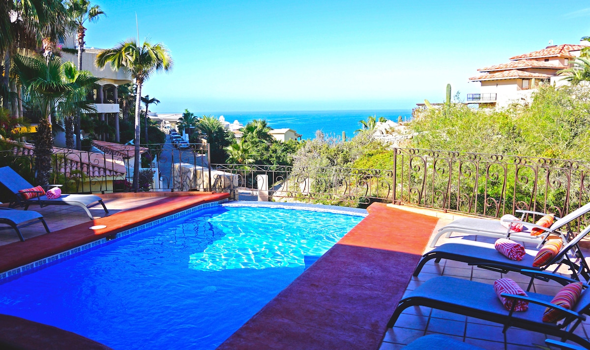A vibrant swimming pool is framed by lounge chairs, set against a backdrop of lush greenery and ocean views. The clear blue water reflects the sunny sky, while the surrounding patio offers a relaxing space to unwind.