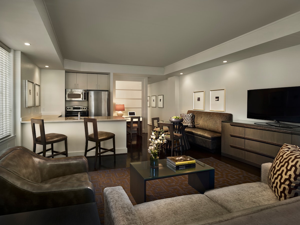 A spacious living area features contemporary furniture, including a sofa and comfortable chairs arranged around a coffee table. A dining table for four can be seen in the background, with a fully equipped kitchen showcasing stainless steel appliances. Natural light filters through the window treatments.