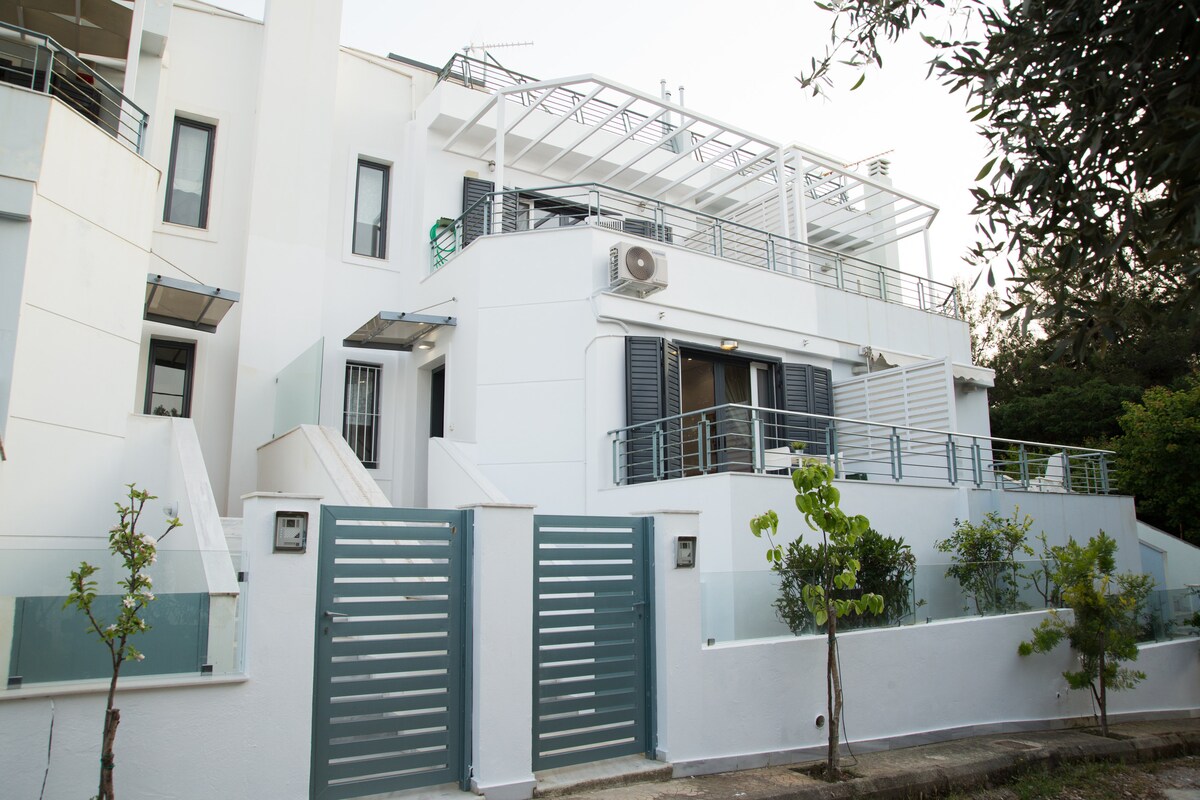 A modern semidetached maisonette is presented with a clean white exterior. The building features a tiered design with balconies and large glass windows. Green shrubs and trees border the entrance, providing a touch of nature in the serene residential area.