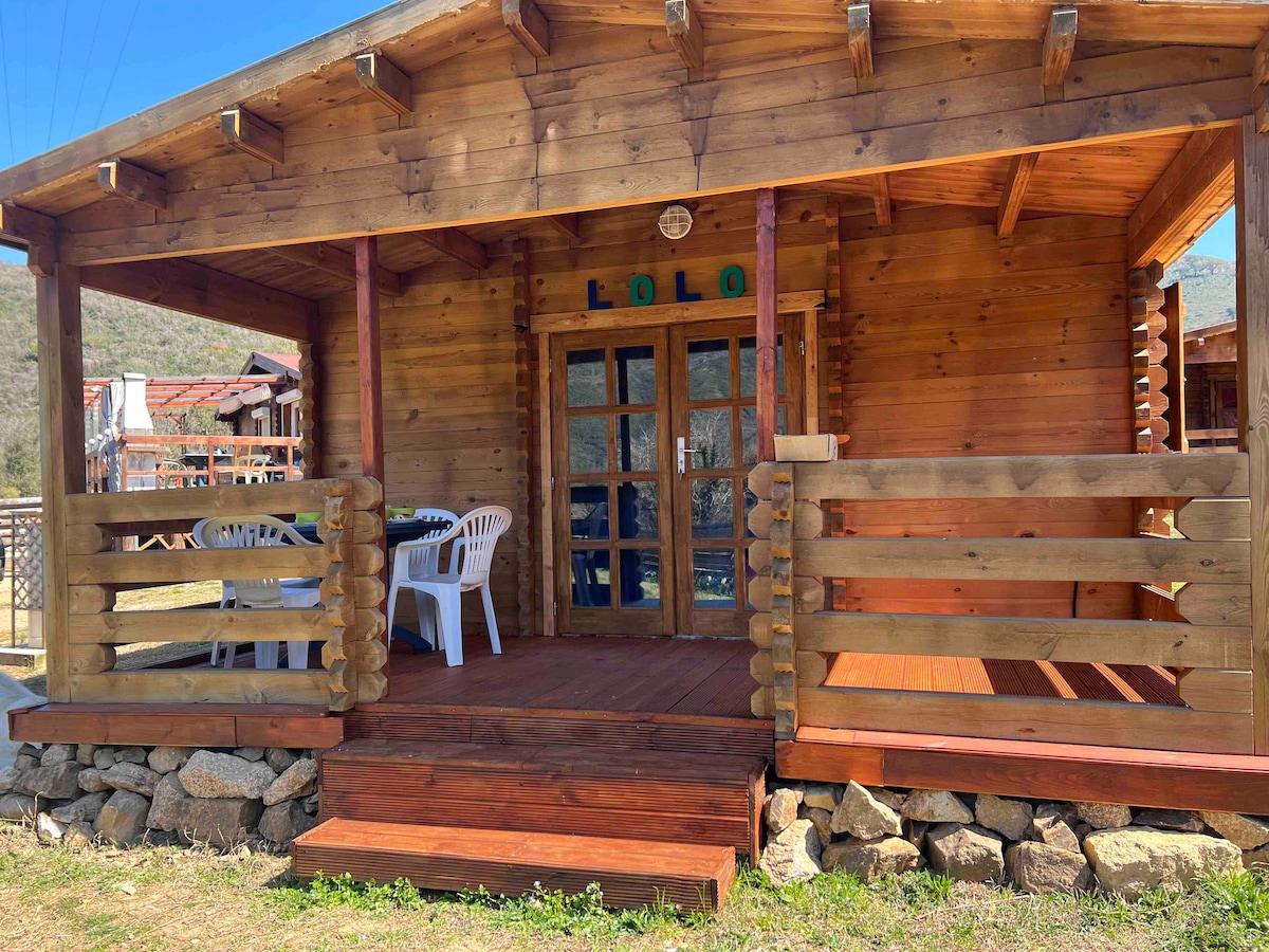 A charming wooden cabin is featured with a welcoming porch. The entryway includes a set of double glass doors and comfortable outdoor seating arranged around a small table. Surrounding greenery adds to the peaceful environment, with a stone border outlined along the base.