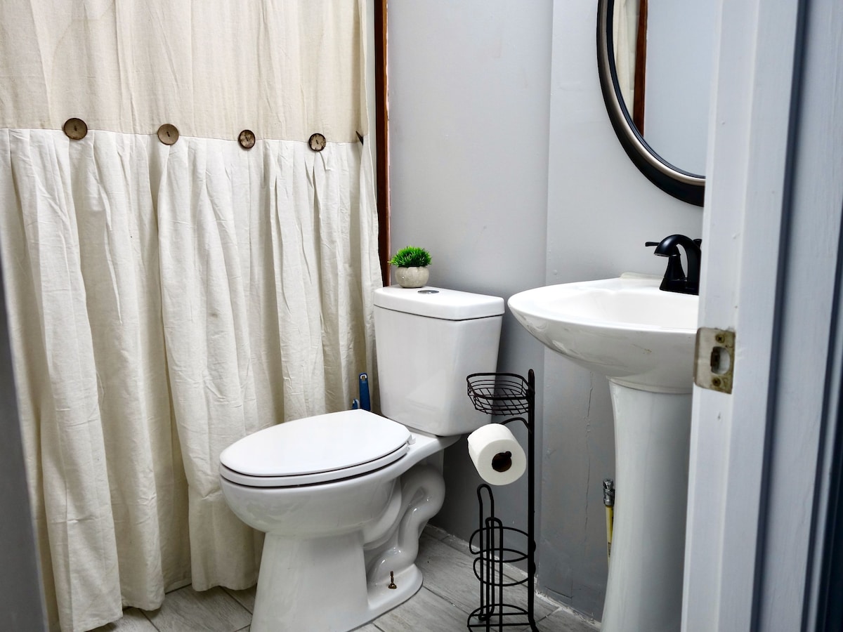 A bathroom is presented, featuring light-colored walls and a round mirror above a white sink. A toilet is positioned adjacent to the sink, while a decorative plant sits on top. A shower curtain in a neutral tone partially conceals the tub area, and a holder with toilet paper is visible.