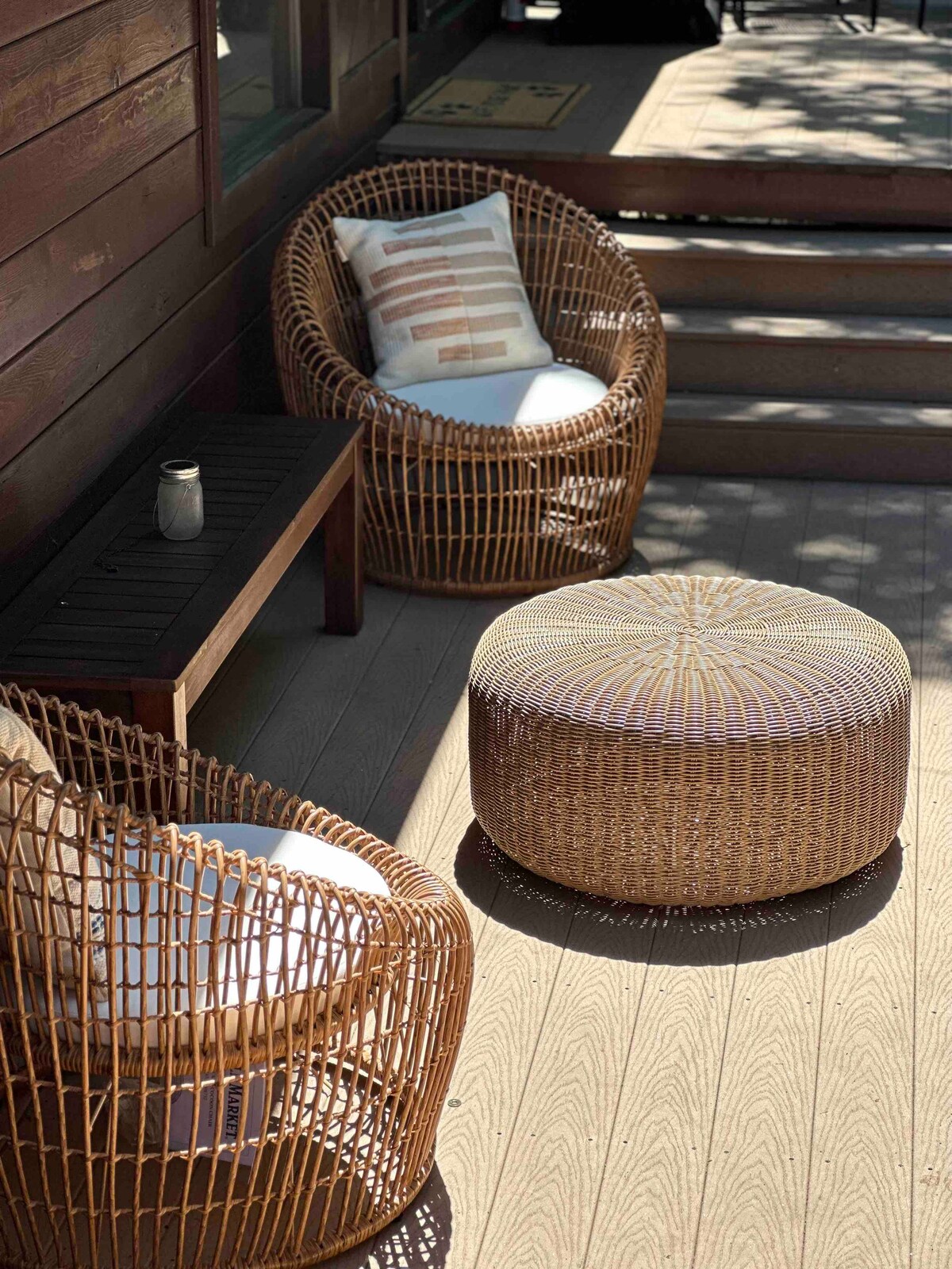 A set of two wicker chairs with cushions and a round ottoman is arranged on the wooden patio. Sunlight filters through the surrounding greenery, casting soft shadows on the deck. A small table sits between the chairs, providing a surface for drinks or snacks.