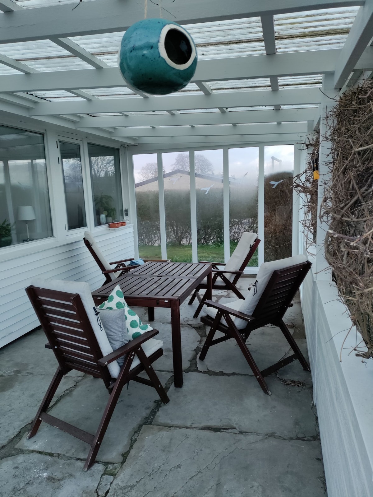 A covered terrace is furnished with a wooden table and four folding chairs. A decorative cushion with a geometric design is featured on one chair. Clear glass doors provide views into the garden, while light filters through the translucent roof.