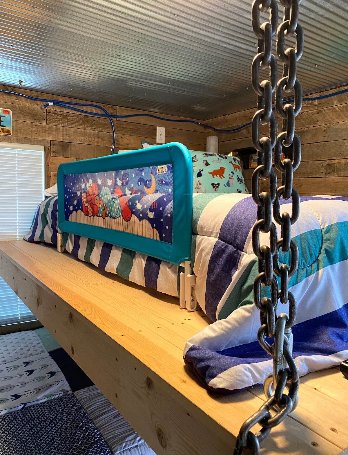 A custom bunk bed is displayed in a cozy sleeping area, featuring a colorful quilt with a playful design. A safety rail is visible along the side of the upper bunk. Textured corrugated walls provide an interesting backdrop, while ambient lighting creates a relaxed atmosphere.