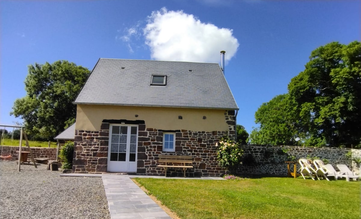 The two-story house features a stone facade combined with a light-colored upper section. A gravel pathway leads to the entrance, framed by a small area of lawn. Large trees and green fields are visible in the background under a clear blue sky.