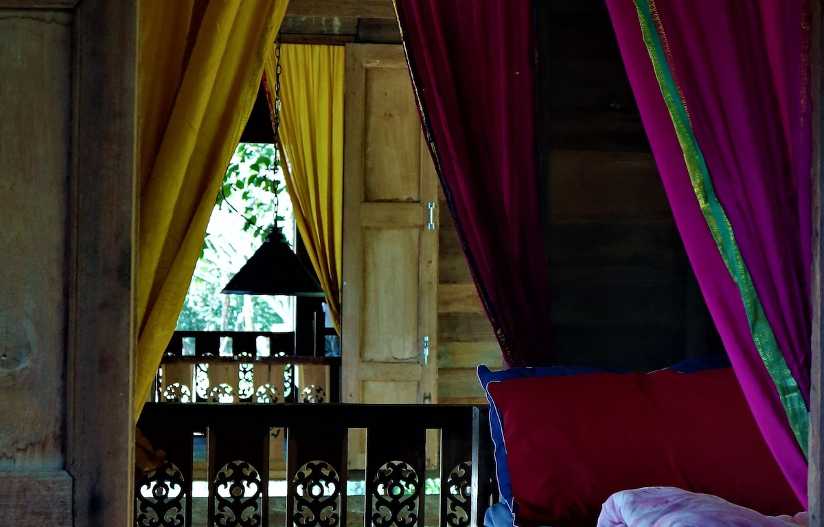 A glimpse into a cozy bedroom is offered, featuring colorful curtains in yellow and pink that frame the view of a wooden door. Natural light filters through, illuminating the rich textures of the wooden interior and highlighting decorative elements in the room.