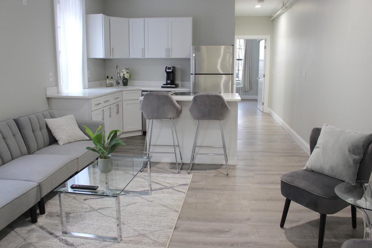 A modern living area features a gray sectional sofa adjacent to a glass coffee table. Two bar stools are positioned at a kitchen counter, with sleek white cabinetry and stainless steel appliances visible in the background. Soft natural light enters through the window.