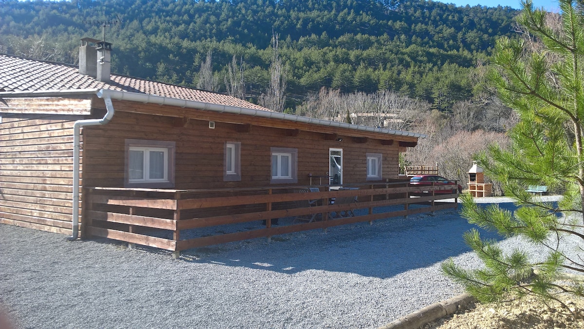 A wooden chalet is set against a mountainous backdrop, featuring a spacious exterior with a gravel driveway and wooden fencing. Multiple windows allow for natural light, and trees are visible in the foreground, contributing to the serene environment.