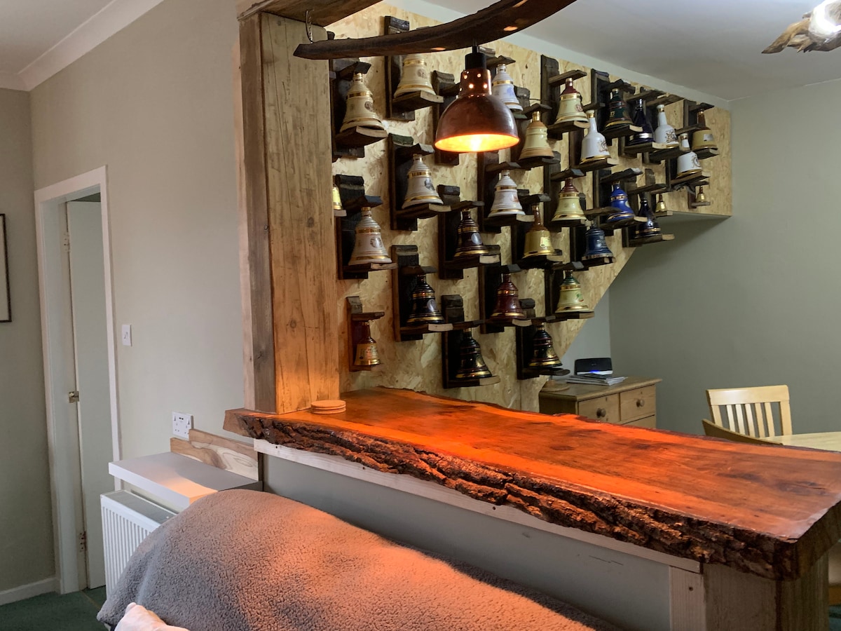 A rustic bar area is highlighted by a unique display of various antique bells mounted on a wooden wall. The bar features a natural wood countertop, and warm lighting creates a cozy ambiance, inviting conversation and relaxation.