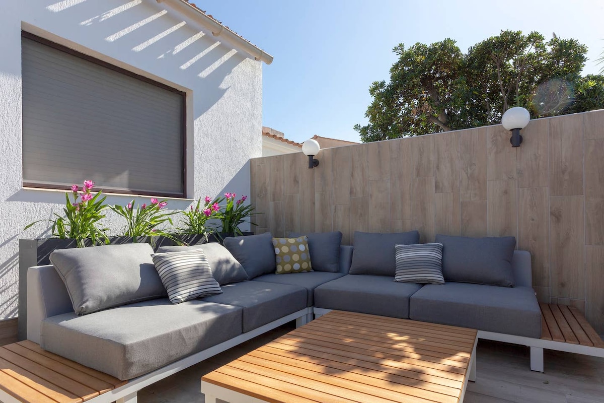 A spacious outdoor seating area features a gray sectional sofa adorned with patterned cushions. A wooden coffee table sits in the center. Lush green plants and vibrant flowers provide a natural backdrop against a light-colored wall, with soft lighting fixtures enhancing the serene environment.