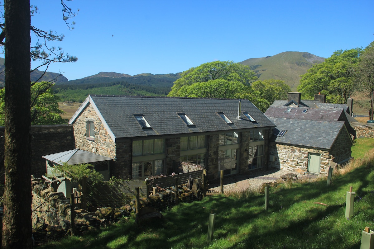 A spacious stone cottage is set against a backdrop of rolling hills and clear blue skies. Large windows allow natural light to flood the interior, while the surrounding greenery adds to the serene landscape.