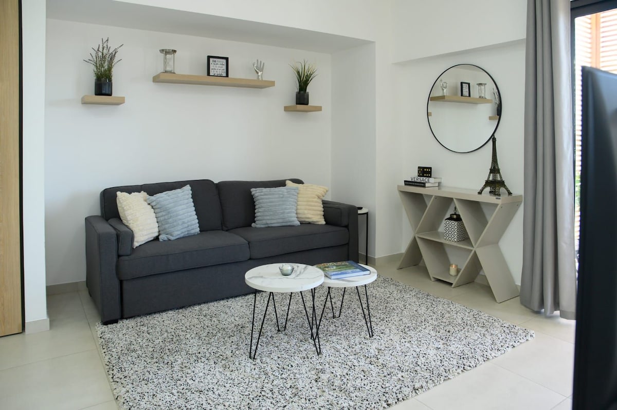 The living area features a gray sofa with decorative pillows, complemented by a round coffee table. A cozy rug anchors the space, while shelving above displays plants and decor. A circular mirror reflects the well-lit environment, creating a sense of openness.