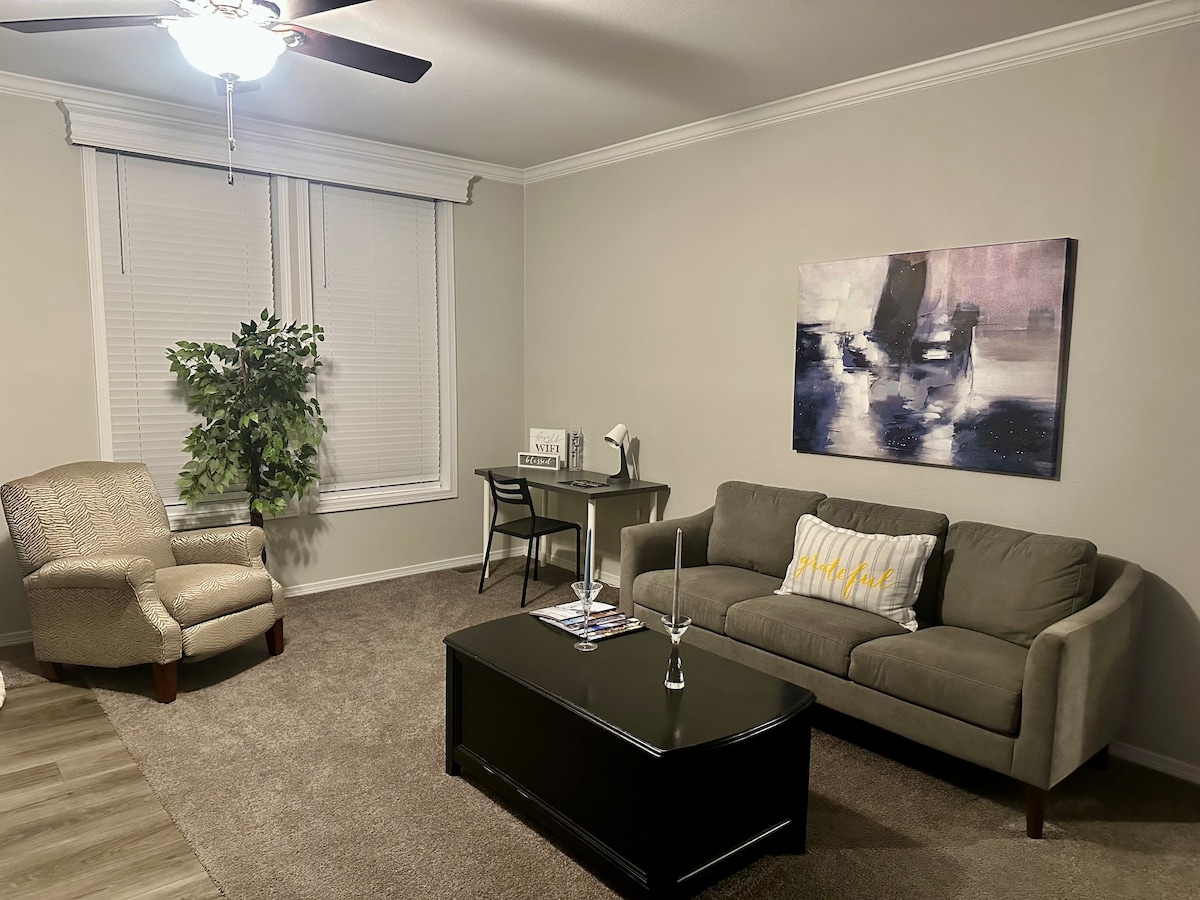 A cozy living area is showcased, featuring a plush gray sofa and a stylish dark coffee table. A soft armchair complements the seating arrangement. Natural light enters through a window adorned with blinds, and a contemporary piece of wall art adds visual interest.