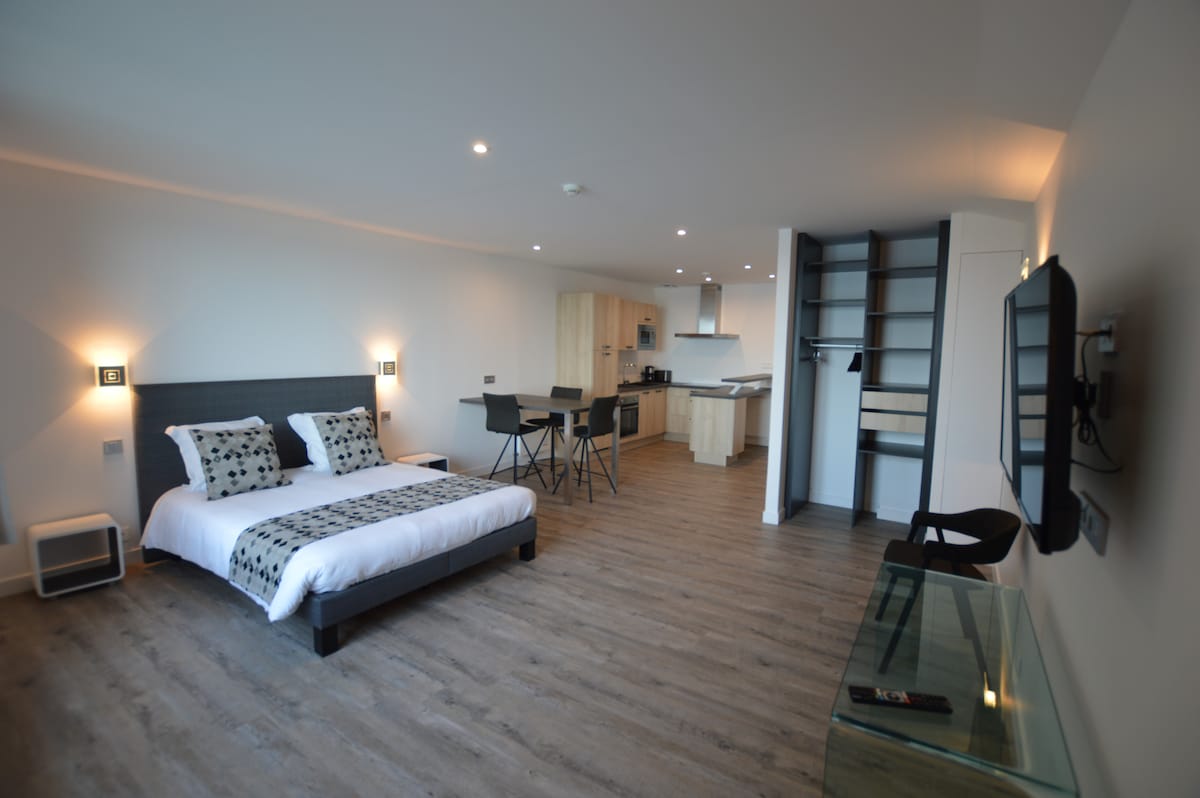 A modern studio layout features a spacious bed with patterned pillows, accompanied by a dining table and chairs. A sleek kitchen area is visible in the background, and a television is mounted on the wall. Warm lighting creates a bright and inviting atmosphere.