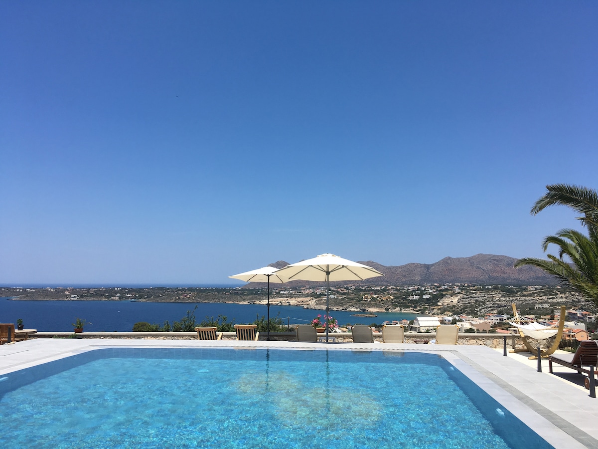 A swimming pool is seen, reflecting clear blue skies and surrounded by lounge chairs and umbrellas. In the background, a panoramic view of the sea and mountains is visible, enhancing the tranquil outdoor space. Lush greenery is present along the pool's edge.