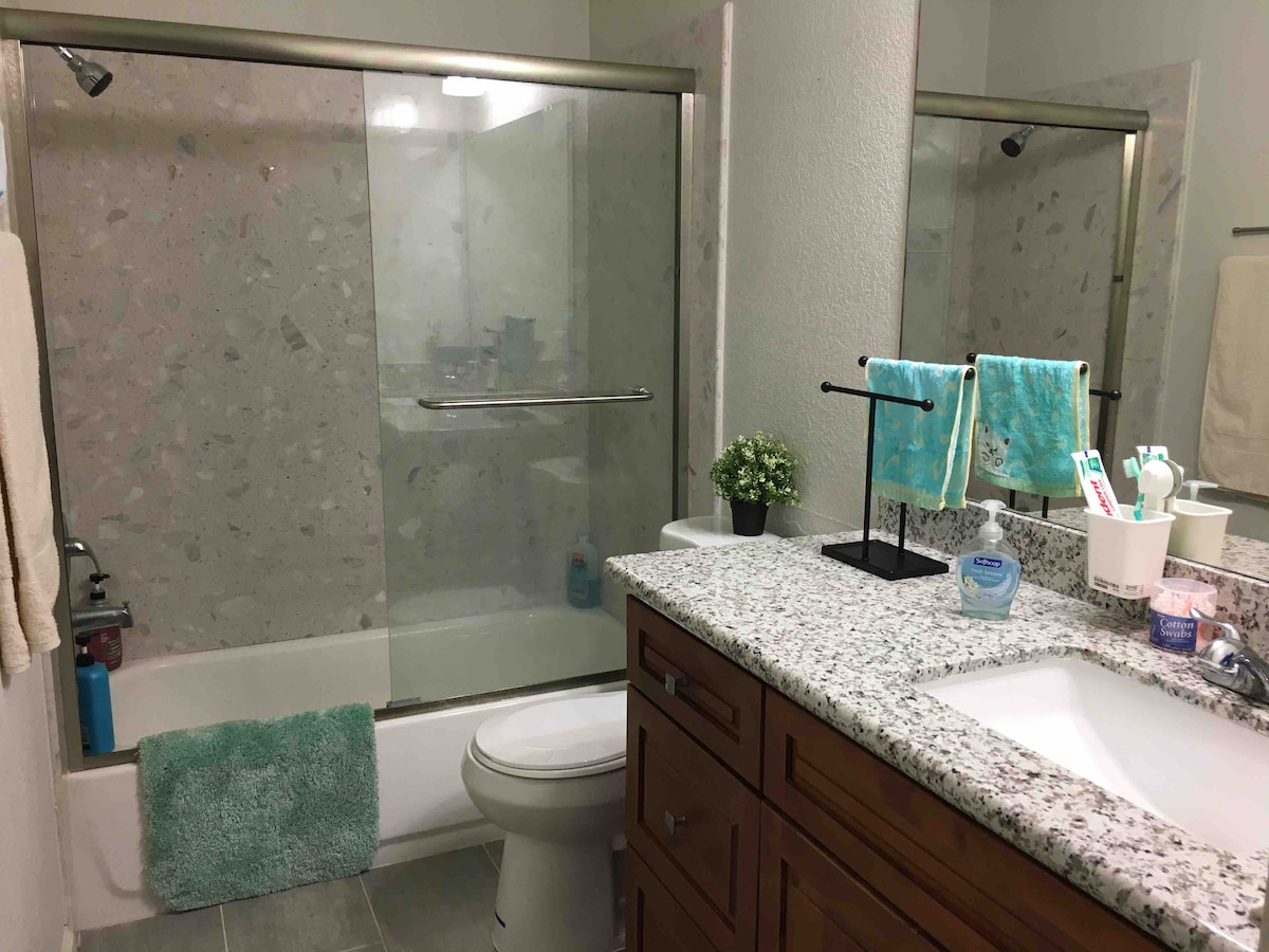 The bathroom features a glass-enclosed shower over a bathtub with granite countertops and a wooden vanity. Complimentary toiletries include a small plant, body lotion, swabs, and a dental kit. A towel bar displays neatly arranged towels, while a light-colored bath mat is positioned in front of the tub.